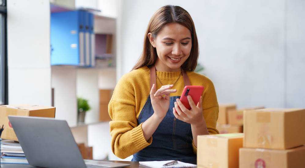 Woman business owner surrounded by boxes and a laptop, smiling at the screen of the cell phone in her hand.