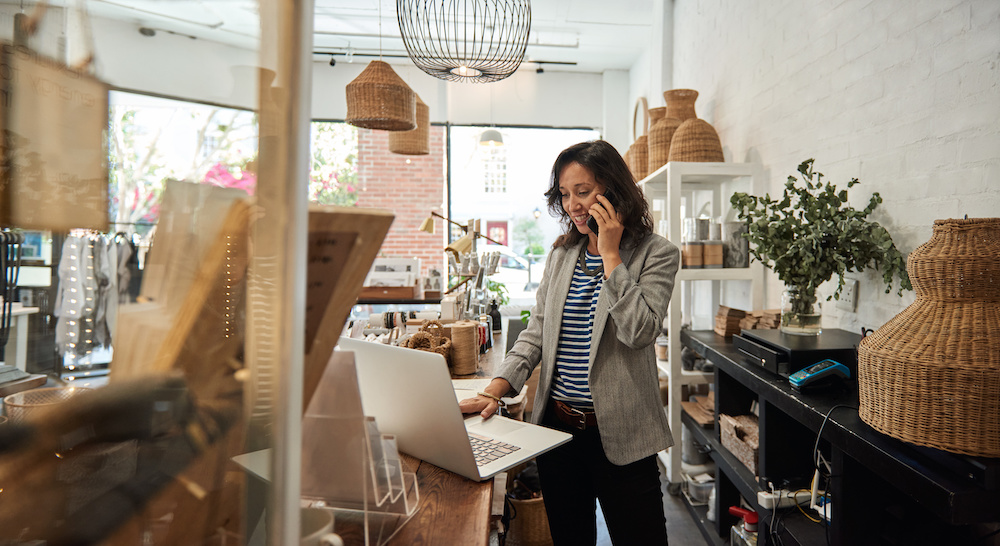 Smiling young woman standing behind a counter in her stylish boutique working on a laptop and talking on a cellphone.