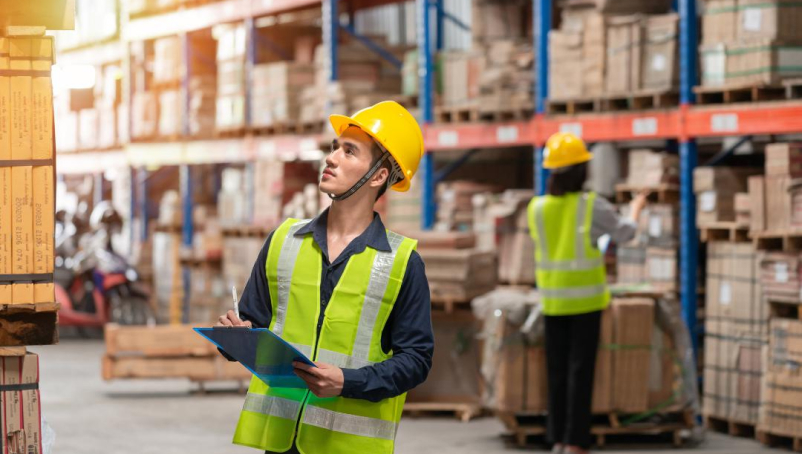 A warehouse technician carrying a clipboard and wearing a reflective vest and a hardhat looks at products in a warehouse.