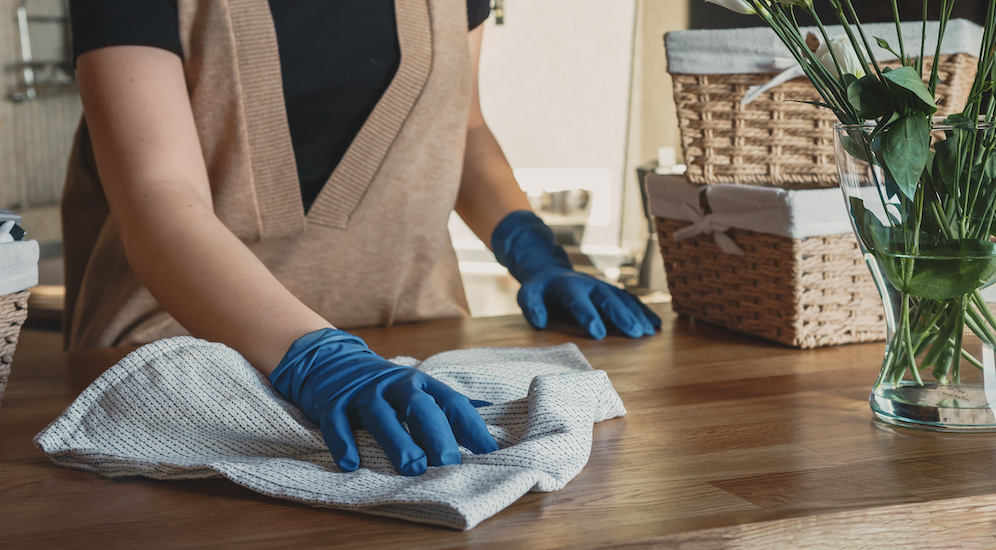 A cleaner wipes a kitchen counter with a cloth while wearing blue gloves.