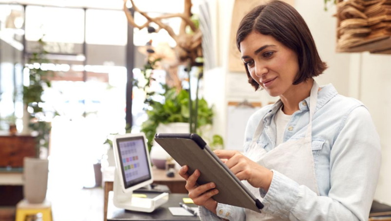 A young woman wearing an apron and using a tablet stands by a counter with a POS system on it.