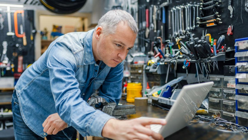 A man, standing in a workroom with handheld tools organized on the walls, uses a laptop.