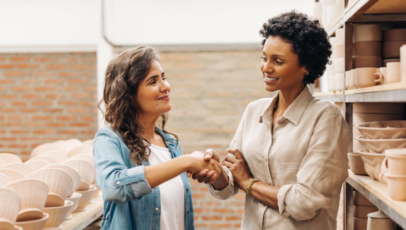 Two women standing in a pottery or ceramics store shake hands.