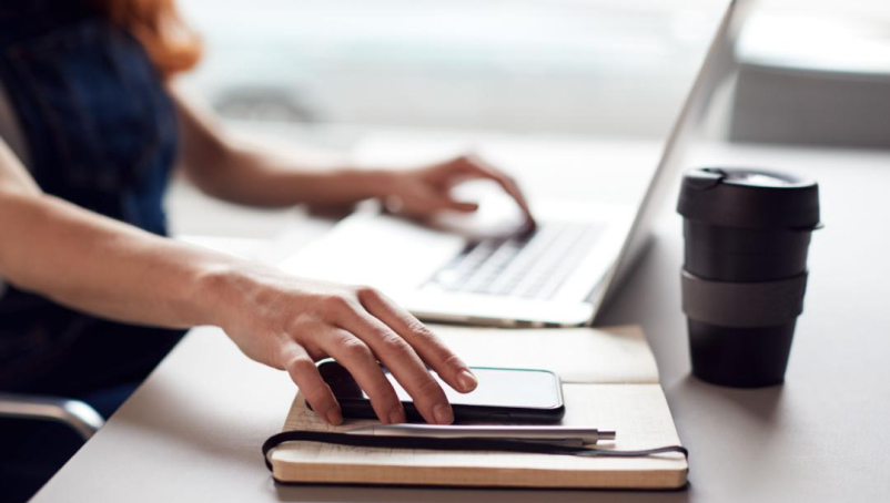 A business owner sits at a desk working on a laptop and reaching for her phone.