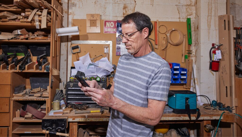 A contractor stands in his workshop, looking at his phone, in front of a workbench and a shelf with tools.